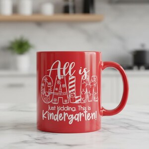 May include: A red ceramic mug with a white handle. The mug features the text "All is Calm, Just kidding. This is Kindergarten!" in a whimsical font. The mug is sitting on a marble surface.