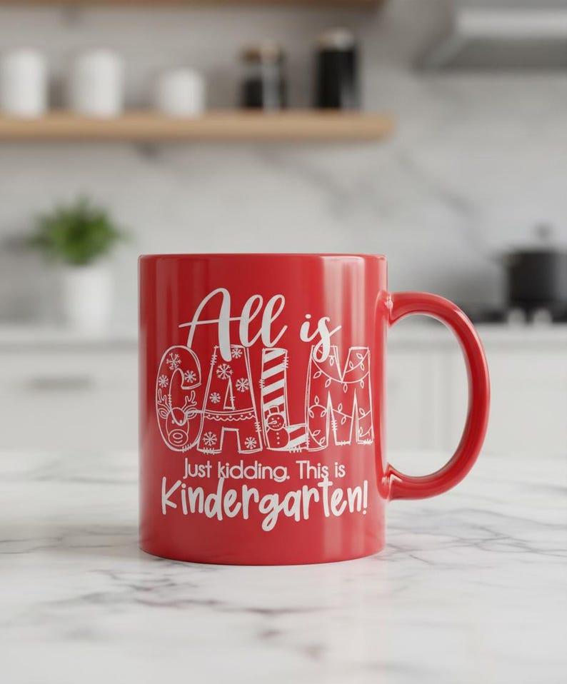 May include: A red ceramic mug with a white handle. The mug features the text "All is Calm, Just kidding. This is Kindergarten!" in a whimsical font. The mug is sitting on a marble surface.