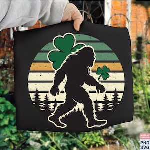 May include: Black t-shirt featuring a Bigfoot silhouette with a green shamrock and a retro sunset design. The design includes a walking Bigfoot, a large shamrock, and a smaller shamrock, with a forest at the bottom. The shirt is held by a person.