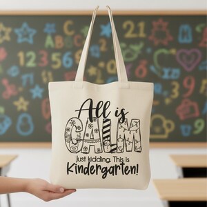May include: Cream-colored tote bag with the text "All is CALM, Just kidding. This is Kindergarten!" in black. The word "CALM" is decorated with snowflakes, stripes, and Christmas lights. The bag has two long handles.