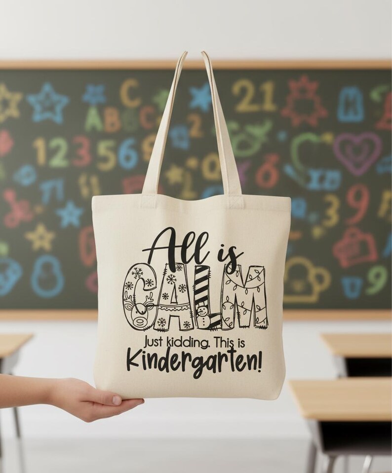 May include: Cream-colored tote bag with the text "All is CALM, Just kidding. This is Kindergarten!" in black. The word "CALM" is decorated with snowflakes, stripes, and Christmas lights. The bag has two long handles.