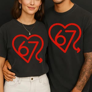 Puede incluir: Dos personas con camisetas grises oscuras a juego. Cada camiseta presenta un dise&ntilde;o de coraz&oacute;n rojo con el n&uacute;mero '67' en su interior. Las camisetas son de estilo casual, adecuadas para el uso diario.