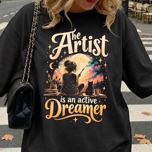 Può includere: Una t-shirt nera con una stampa grafica di una persona seduta a dipingere davanti a una grande luna. Il testo recita The Artist is an active Dreamer. L'illustrazione include pennelli, un gatto e una tazza su una superficie.