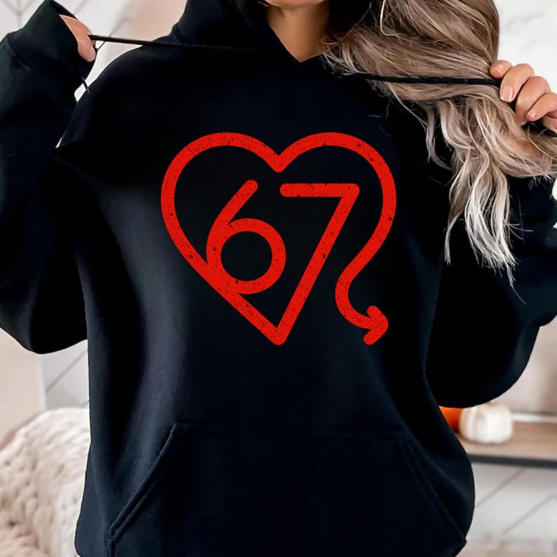 Puede incluir: Sudadera con capucha negra con un gr&aacute;fico rojo en forma de coraz&oacute;n. El gr&aacute;fico presenta el n&uacute;mero "67" en una fuente estilizada. La sudadera tiene un bolsillo delantero y una capucha con cord&oacute;n.