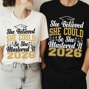 Può includere: Due magliette, una bianca e una nera, con la scritta "She Believed SHE COULD So She Mastered It 2026". Il design include un cappello di laurea, stelle e pergamene, celebrando la classe del 2026.