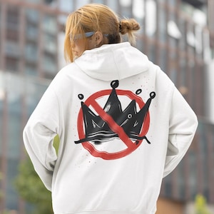 May include: White hoodie with a graphic on the back featuring a black crown crossed out by a red circle. The hoodie has a drawstring hood and long sleeves. The background is a blurred outdoor scene.