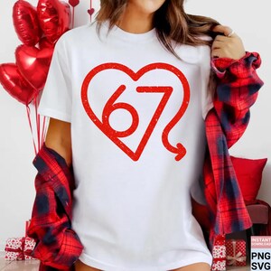 Puede incluir: Camiseta blanca con un gran dise&ntilde;o de coraz&oacute;n rojo. Dentro del coraz&oacute;n est&aacute; el n&uacute;mero 67 con una cola curva. La camiseta forma parte de un tema de San Valent&iacute;n, con globos rojos en forma de coraz&oacute;n en el fondo.