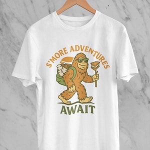 Puede incluir: Camiseta blanca con un dibujo de Bigfoot con gafas de sol, una mochila y un s'more en un palo. El texto "S'MORE ADVENTURES AWAIT" está impreso encima y debajo de la ilustración de Bigfoot en naranja y verde.