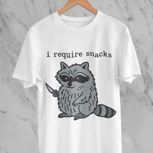 May include: White t-shirt featuring a cartoon raccoon holding a knife, with the text "i require snacks" above the raccoon. The raccoon is gray and black, with a striped tail. The shirt is hanging on a wooden hanger.