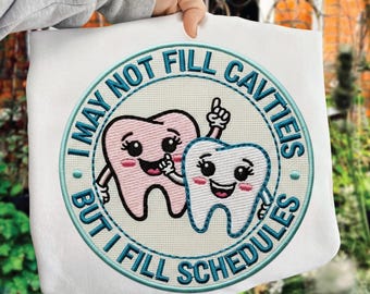 Op de afbeelding: Wit sweatshirt met een geborduurd ontwerp van twee cartoon tanden met lachende gezichten. Het ontwerp is omcirkeld door turquoise tekst die luidt: "I MAY NOT FILL CAVITIES BUT I FILL SCHEDULES."