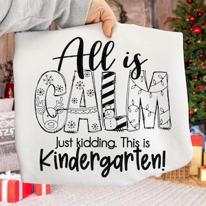 May include: White sweatshirt with the words "All is CALM" in black script, with the letters decorated with snowflakes, a reindeer, a snowman, and Christmas lights. Below, the text reads "Just kidding. This is Kindergarten!"