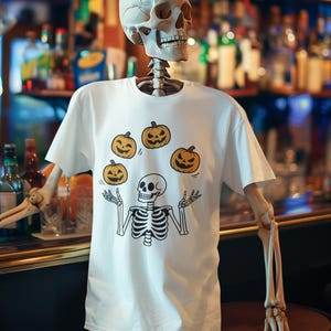 Puede incluir: Una camiseta blanca con un esqueleto de dibujos animados haciendo malabarismos con calabazas. El diseño muestra un esqueleto con un cráneo y una caja torácica, y las calabazas tienen caras sonrientes. La camiseta la lleva un modelo de esqueleto en un bar.