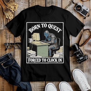 May include: Black t-shirt with a graphic of a knight in armor at a computer, with the text "BORN TO QUEST" above and "FORCED TO CLOCK IN" below. The knight is sitting at a desk with a vintage computer and stacks of papers.