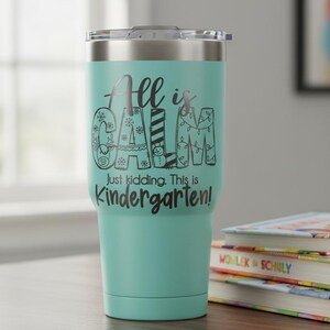 May include: A teal insulated tumbler with a silver lid. The tumbler has the text "All is Calm" in a decorative font, with the words "Just kidding. This is Kindergarten!" below. The tumbler is sitting on a wooden surface next to a stack of books.
