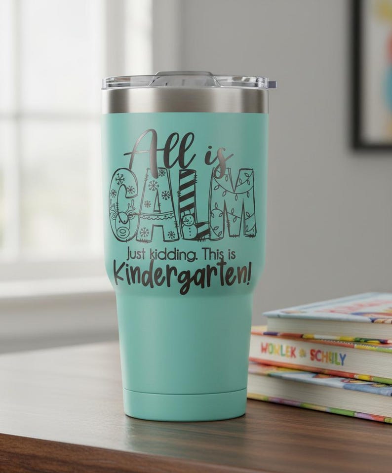 May include: A teal insulated tumbler with a silver lid. The tumbler has the text "All is Calm" in a decorative font, with the words "Just kidding. This is Kindergarten!" below. The tumbler is sitting on a wooden surface next to a stack of books.