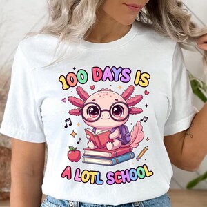May include: White t-shirt with a colorful graphic featuring an axolotl wearing glasses and reading a book. The text reads "100 DAYS IS A LOTL SCHOOL" in various colors. An apple, pencil, and musical notes are also included.