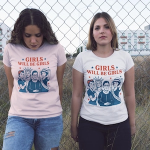May include: Two women wearing t-shirts with the text "GIRLS WILL BE GIRLS" and an illustration of notable women. One shirt is light pink, the other is white. They stand in front of a chain-link fence.