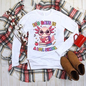 May include: White long-sleeve shirt with a colorful graphic of an axolotl wearing glasses and reading a book. The text on the shirt reads "100 Days is A LOTL School". The shirt is displayed on a plaid blanket.