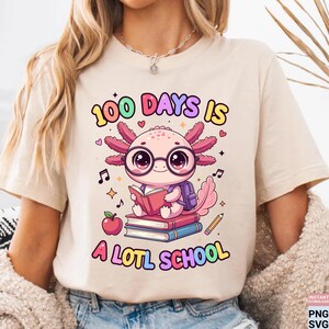 May include: Beige t-shirt with a colorful graphic featuring an axolotl wearing glasses and reading a book. The text reads "100 DAYS IS A LOTL SCHOOL" in a playful font, with musical notes and an apple.