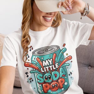 May include: White t-shirt featuring a cartoon soda can design with the text "MY LITTLE SODA POP". The can is teal with red and orange accents, and the design includes musical notes and cartoon faces. A beige cap is worn.
