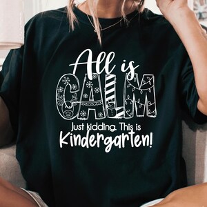 May include: Black t-shirt with the words "All is CALM" in white, with snowflake and holiday-themed designs incorporated into the letters. Below, the text reads "Just kidding. This is Kindergarten!"