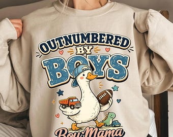 May include: Beige sweatshirt with the text "OUTNUMBERED BY BOYS" in blue and yellow. A cartoon goose wearing a blue cap and holding a football and a toy truck is featured. The text "Boy Mama AND I LOVE IT" is also included.