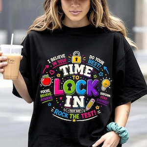 Puede incluir: Camiseta negra con un diseño gráfico colorido. El diseño incluye el texto "Time to Lock In" con una manzana, una bombilla y un lápiz. La camiseta también tiene las frases "I Believe in You!" y "Do Your Best!"