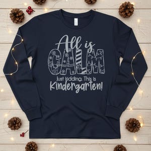 May include: Navy blue long-sleeve shirt with the text "All is CALM, Just kidding. This is Kindergarten!" in a whimsical font. The word "CALM" is decorated with snowflakes, a reindeer, a snowman, and Christmas lights.