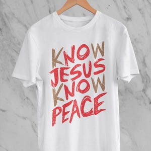 May include: White t-shirt with the words "KNOW JESUS KNOW PEACE" in red and brown lettering. The shirt is hanging on a wooden hanger against a white and gray marble background.