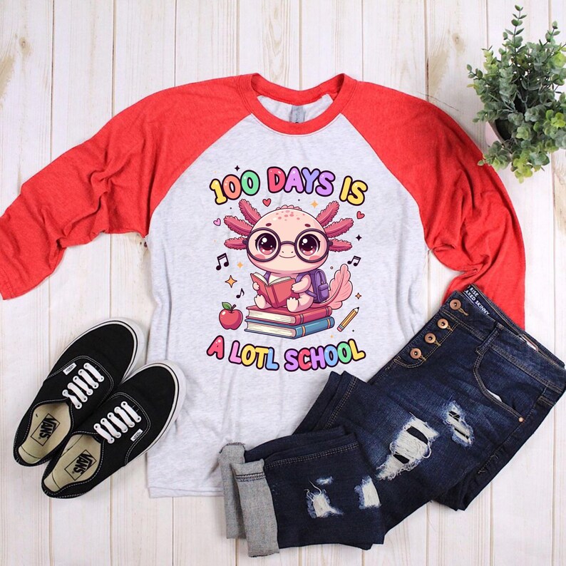 May include: A gray and red raglan shirt with the text "100 DAYS IS A LOTL SCHOOL" and a cartoon axolotl wearing glasses and reading a book. The image also includes black shoes and blue jeans.