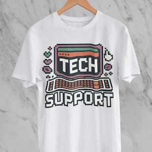 Könnte beinhalten: Weißes T-Shirt mit einem pixeligen Computerdesign. Das Design zeigt die Worte "TECH SUPPORT" in einer Retro-Schriftart, mit einer Tastatur und einer Bildschirmgrafik. Das T-Shirt hängt an einem Holzbügel.