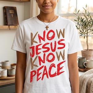 May include: A white short-sleeved t-shirt featuring the text "KNOW JESUS KNOW PEACE" printed in a distressed, bold red font. The shirt is worn with a simple brown wooden cross pendant on a thin cord necklace, resting against the chest area.