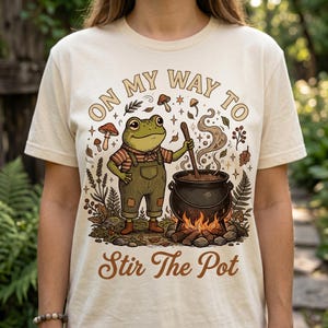 Op de afbeelding: Crèmekleurig T-shirt met een cartoonkikker in een overall die een ketel boven een vuur roert. De tekst "ON MY WAY TO" en "Stir The Pot" zijn boven en onder de kikkerillustratie gebogen. Het ontwerp bevat paddenstoelen en bladeren.