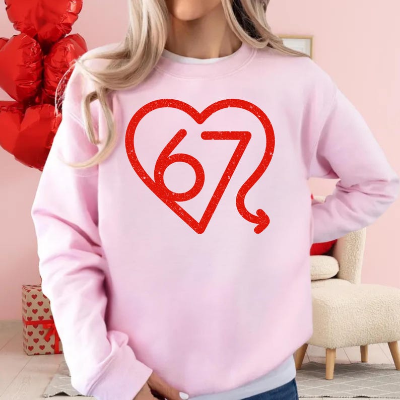 Puede incluir: Sudadera rosa claro con un dise&ntilde;o de coraz&oacute;n rojo. Dentro del coraz&oacute;n est&aacute; el n&uacute;mero 67 en una fuente estilizada. La sudadera es de color liso y tiene mangas largas.