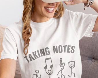 May include: White t-shirt with the text "TAKING NOTES" in gray, along with stick figure illustrations of musical notes and people. A beige baseball cap is worn. The shirt is a casual, music-themed apparel item.