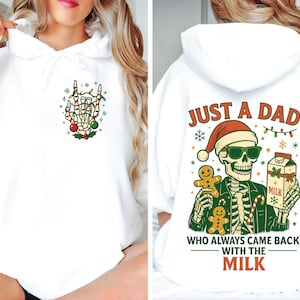 May include: White hoodie with a Christmas-themed skeleton graphic. The front features a skeleton hand making a rock-and-roll gesture, decorated with Christmas lights and ornaments. The back displays a skeleton wearing a Santa hat, holding milk and gingerbread men, with the text "JUST A DAD WHO ALWAYS CAME BACK WITH THE MILK."