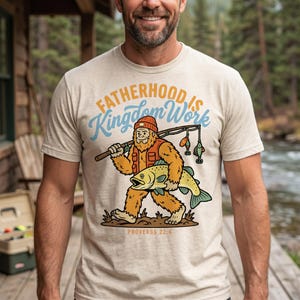 May include: A heathered beige t-shirt featuring a graphic of a cartoon Bigfoot wearing a beanie and vest, holding a fishing rod and a large fish. The text reads "FATHERHOOD IS Kingdom Work" in orange and blue, with "PROVERBS 22:6" below.
