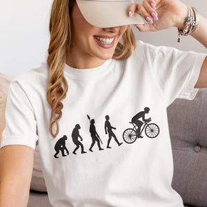 Puede incluir: Camiseta blanca con un gráfico negro que representa la evolución humana hacia un ciclista. El diseño muestra una progresión de simio a ciclista. La persona de la imagen lleva una gorra de béisbol beige.