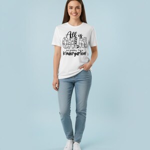 May include: A white t-shirt with the text "All is Calm, Just kidding, This is Kindergarten!" in black. The person is wearing blue jeans and white sneakers. The background is a light blue.