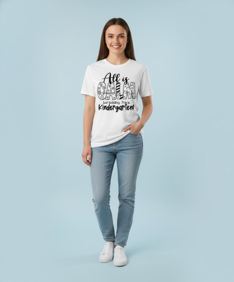 May include: A white t-shirt with the text "All is Calm, Just kidding, This is Kindergarten!" in black. The person is wearing blue jeans and white sneakers. The background is a light blue.