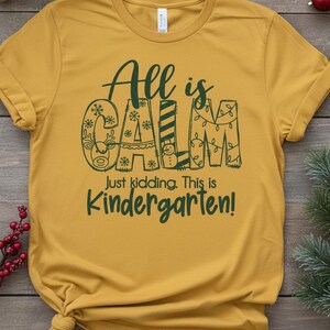May include: Mustard yellow t-shirt with the text "All is CALM" in dark green, with the letters decorated with snowflakes, a reindeer, and Christmas lights. Below, it reads "Just kidding. This is Kindergarten!"