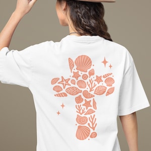 May include: White t-shirt featuring a coral-colored cross design composed of seashells, starfish, and coral. The design is on the back of the shirt. The person is wearing a brown hat.