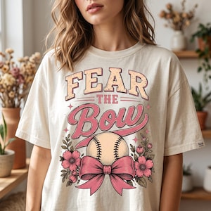 以下が含まれることがあります： ピンクとベージュの文字で「FEAR THE Bow」と書かれたグラフィックプリントが特徴のクリーム色のTシャツ。中央には野球ボールのイラストが描かれ、大きなピンクのリボンとピンクの花の要素、緑の葉が添えられています。