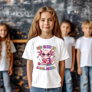 May include: A white t-shirt featuring a colorful cartoon axolotl wearing glasses and reading a book. The text "100 DAYS IS A LOTL SCHOOL" is printed above and below the axolotl illustration. The shirt is worn by a young person.