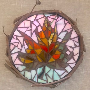 May include: A mosaic art piece of a colorful fall leaf framed with twigs. The leaf is made of small pieces of glass in shades of pink, orange, red, brown, and green.