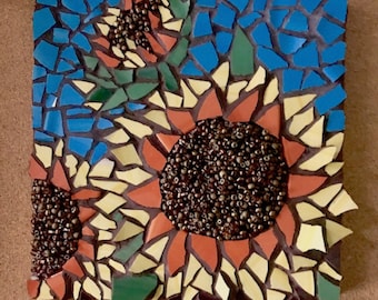 May include: A mosaic artwork featuring three sunflowers with yellow petals, orange centers, and brown seeds. The background is a blue and green mosaic.