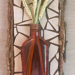May include: A brown glass bottle with a white ceramic tile frame and dried wheat stalks. The bottle is sitting on a white tile background.