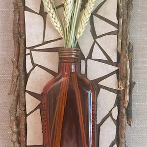 May include: A brown glass bottle with a narrow neck and a white ceramic tile frame. The bottle is filled with dried wheat stalks and is set against a white background.