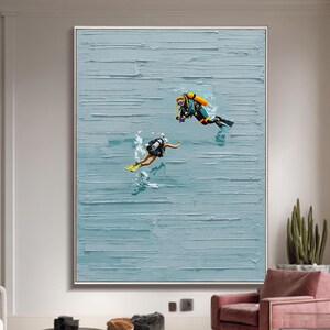Puede incluir: Gran pintura abstracta enmarcada con dos buceadores en el océano. La obra de arte tiene un fondo azul claro texturizado con pinceladas horizontales. Los buceadores llevan equipo negro y amarillo.