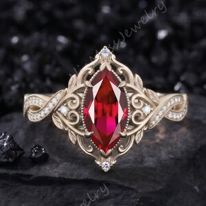 Può includere: Un anello in oro con una gemma rossa centrale taglio marquise, circondata da un'intricata montatura in filigrana con piccole pietre chiare incastonate lungo la fascia. Il design presenta dettagliati motivi a volute e a foglia.